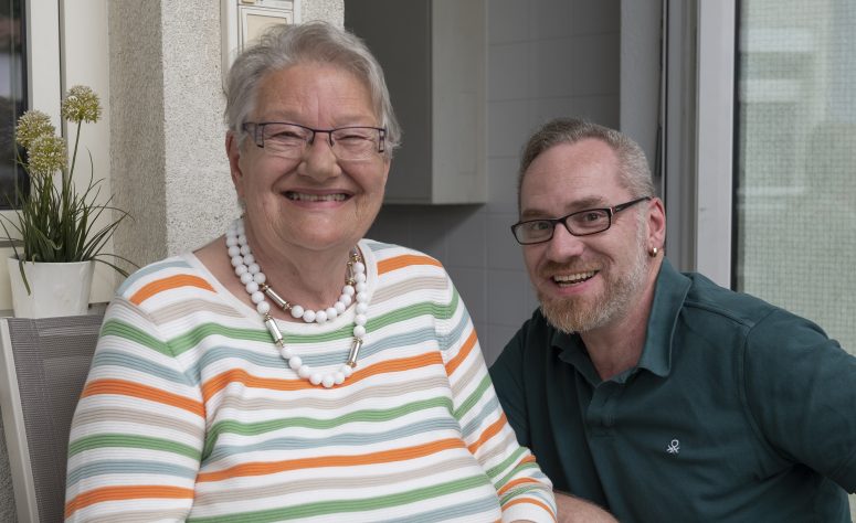 Ältere Dame sitzt draussen lachend mit ihrem Pfleger 